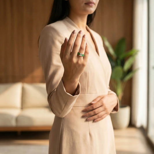Woman in a beige outfit holding her hand to her chest with a blurred indoor background and showing a green ring