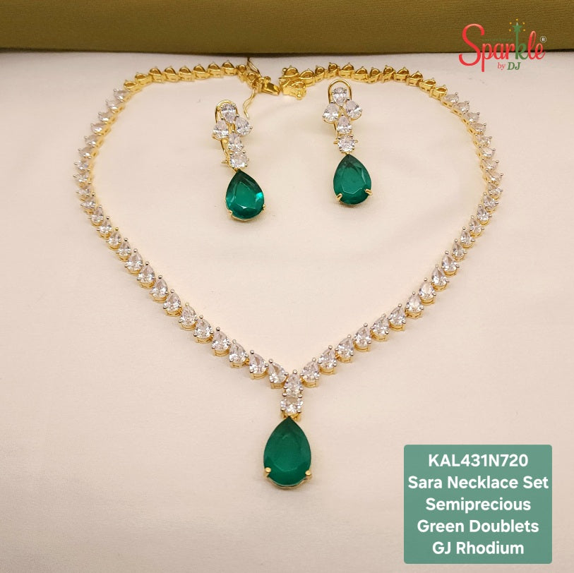 Gold jewelry set featuring a necklace and matching earrings with green teardrop-shaped doublets and clear stones, displayed on a cream surface with a green box edge in the background. A green text box at the bottom right lists the product details.