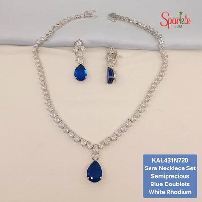 Sara necklace set featuring a silver-tone rhodium chain necklace with clear cubic zirconia stones and a large blue teardrop doublet pendant, accompanied by matching drop earrings. The set is displayed on a beige surface with a white background, featuring a brand logo 'Sparkle by Dr' and a product code label in the corners.