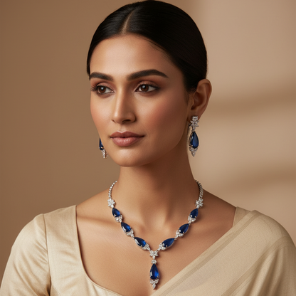 A woman wearing a matching jewelry set featuring a V-neck necklace and drop earrings with deep blue teardrop gemstones and silver-toned pavé settings, paired with a beige top against a warm beige background.