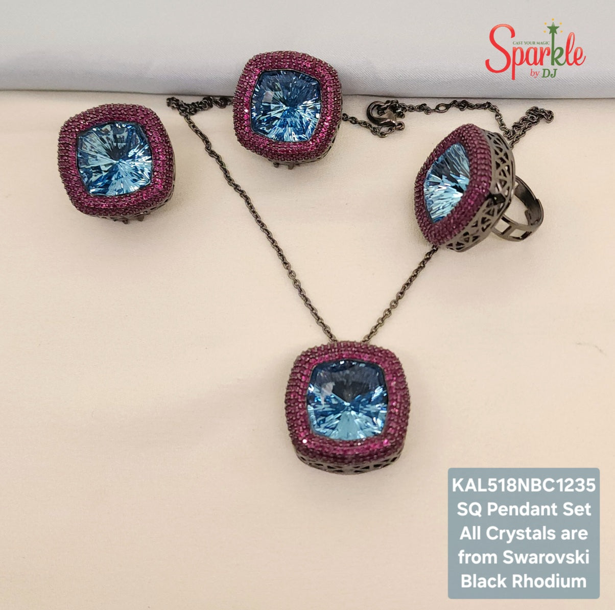 A jewelry set displayed on a light beige surface, including a pendant necklace, two matching earrings, and a ring. The set features large square-cut light blue crystals surrounded by a border of pink stones, with black rhodium metal settings and chains.