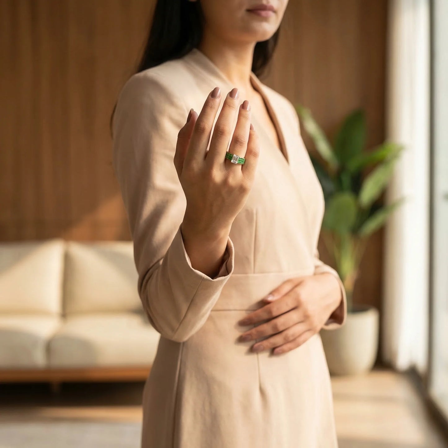Woman in a beige outfit holding her hand to her chest with a blurred indoor background and showing a green ring